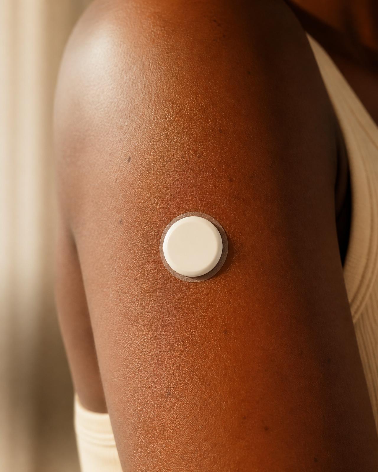 A small circular femmli biosensor worn on the back of a woman's upper arm, similar to a continuous glucose monitor
